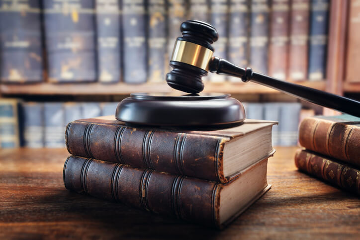 Judge’s gavel resting on a stack of old law books on a wooden desk, with shelves of legal books blurred in the background.
