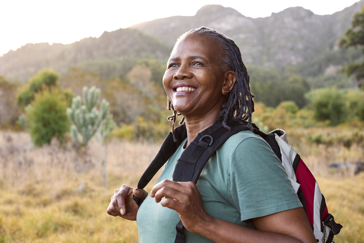 A woman wearing a green shirt and backpack smiles while standing in a grassy landscape with mountains in the background.