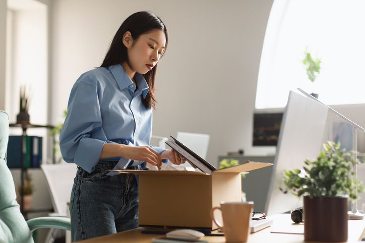 Sad unemployed Asian woman packing personal belongings in cardboard box standing at table desk, feeling stressed and depressed losing her job. Upset dismissed lady leaving office after being fired
