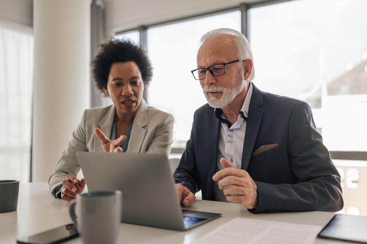 Female executive discussing with senior professional while working on laptop at desk in office