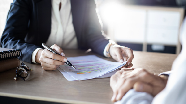 Person in a business suit pointing to highlighted sections of a document during a discussion across a desk, suggesting a contract or legal review.