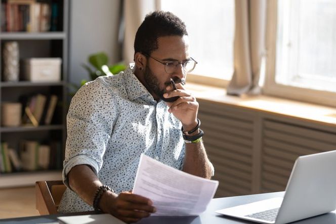 Focused serious african american businessman accountant analyst holding documents looking at laptop computer screen doing online trade market tech research thinking working sit at home office desk