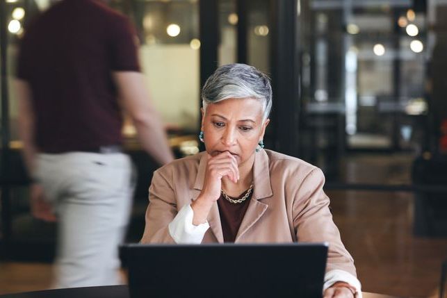 An older person with short gray hair sits at a desk, looking thoughtfully at a laptop screen. A person walks by in the background.