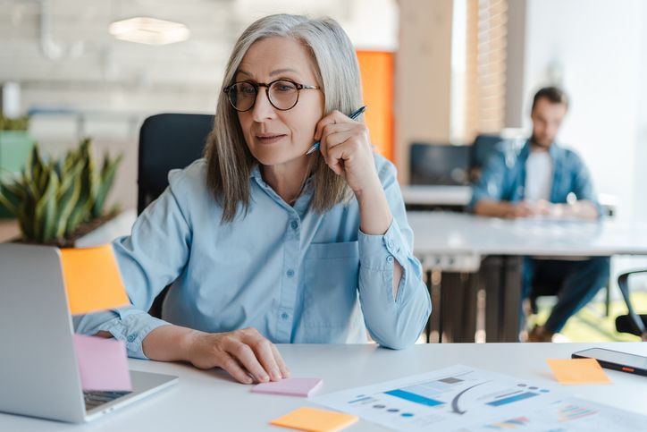 Smiling confident senior businesswoman using laptop, sticky notes looking at monitor in modern office. Gray haired manager wearing stylish eyeglasses sitting at workplace. Scrum, agile concept