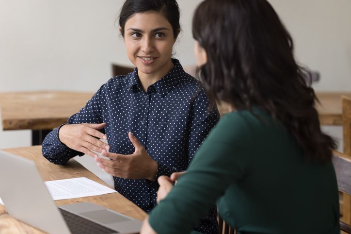 Women colleagues discuss joint task seated at desk with laptop stock