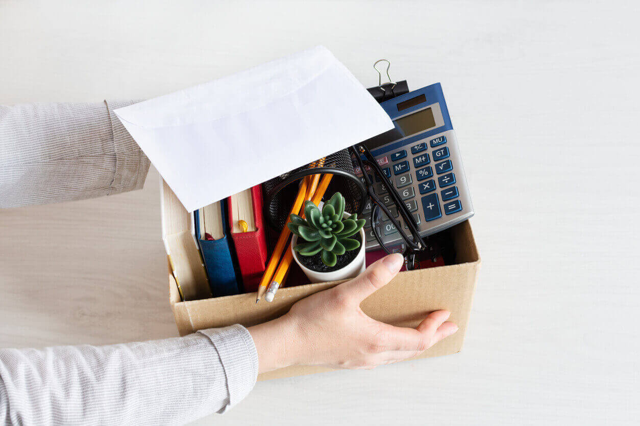 Person holding a cardboard box filled with office supplies, including books, a calculator, pencils, a potted plant, and an envelope, symbolizing job termination or employee furlough.