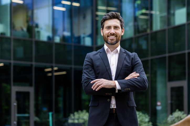 Portrait of mature adult businessman, senior boss with beard smiling and looking at camera, experienced and successful man in business suit outside office building with crossed arms.