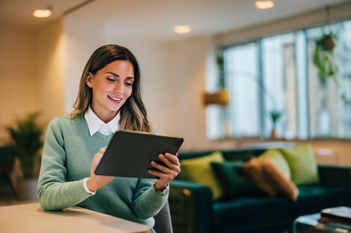 Woman in a green sweater sits at a table, using a tablet in a bright, modern living room.