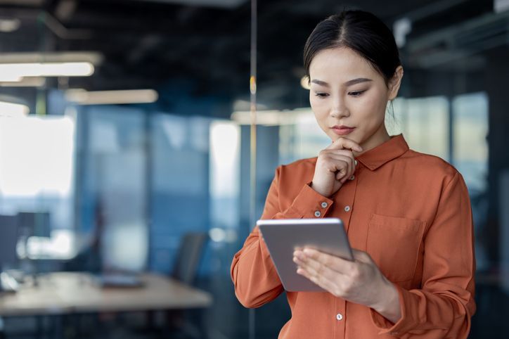 A person stands in an office, looking thoughtfully at a tablet.