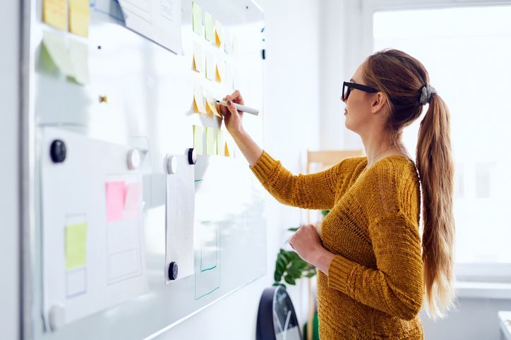 Woman in glasses writing on a whiteboard with sticky notes in a bright office environment.