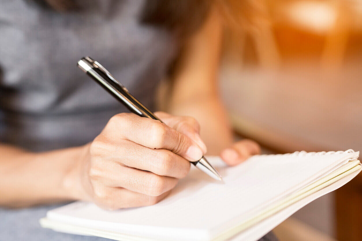 Close-up of a person writing in a spiral notebook with a black pen, sitting indoors with a blurred background.