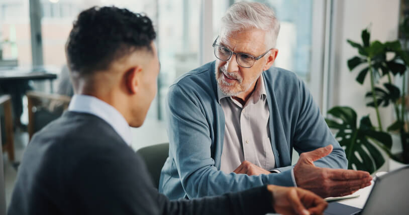 Older man with gray hair and glasses speaking and gesturing during a one-on-one discussion with a younger colleague in a modern office setting.
